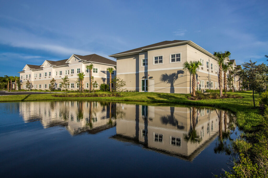 Heritage Oaks at Englewood beige buildings with brown roofs in front of a green lawn with palm trees by the lake.