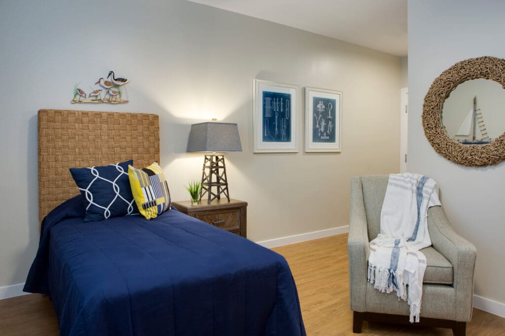 A bedroom with a twin-sized bed with blue bedding and throw pillows, a bedside table with a lamp, and a gray sofa chair with a white throw blanket on it.