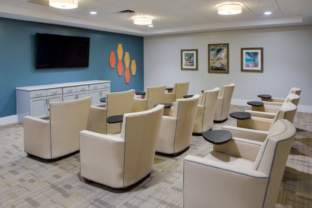 A movie theater room with three rows of beige sofa chairs facing a TV screen at Heritage Oaks of Englewood, a senior living community near Venice, FL.