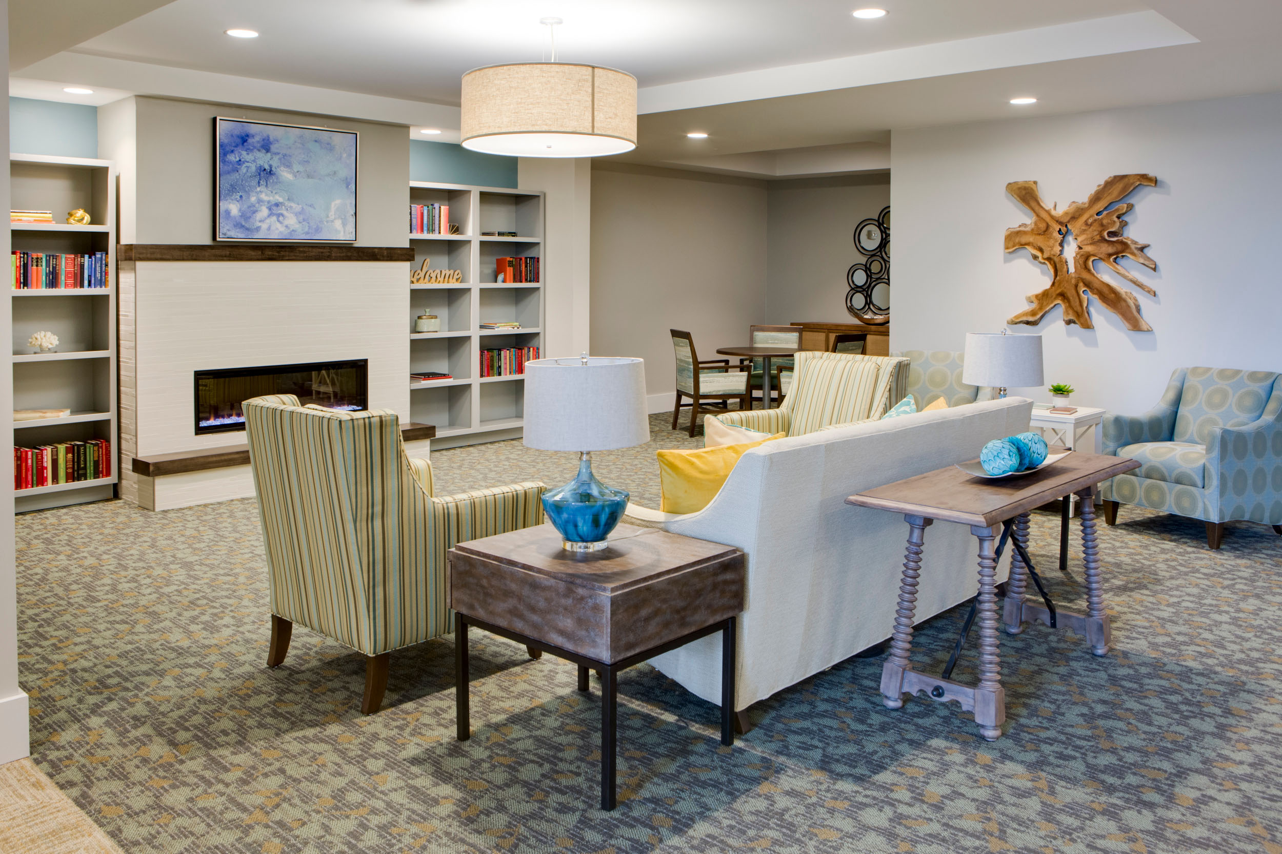 A living room are with a white couch and throw pillows on it, sofa chairs next to it with side tables and lamps on it, located across a digital fireplace that's underneath a wall painting, which is next to two bookshelves with books on them at Heritage Oaks of Englewood, a senior living community near Venice, FL.