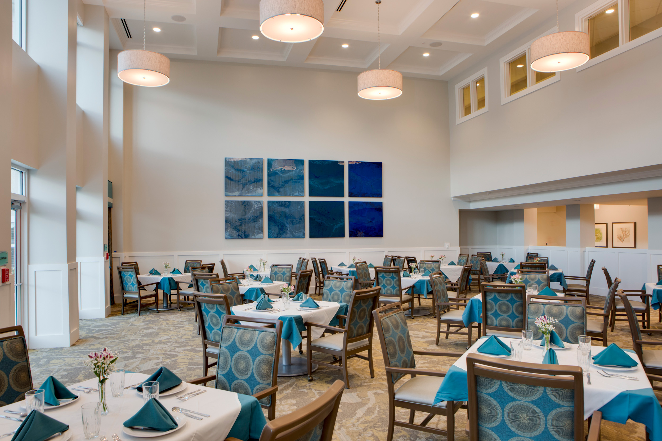 A dining area with square tables covered in blue and white table cloths with plates, glasses, silverware, and blue napkins on them, surrounded by blue and brown patterned chairs at Heritage Oaks of Englewood, a senior living community near North Port, FL.