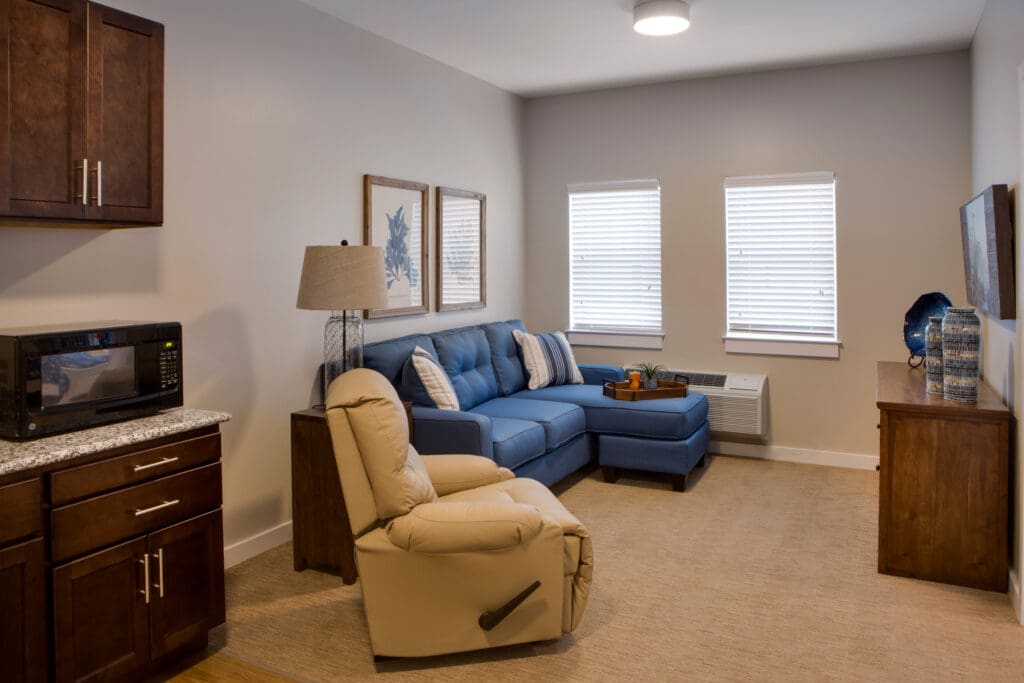A living room with a blue couch facing the TV that's mounted to the wall underneath a dresses, with a mustard-colored reclining sofa chair next to the couch.