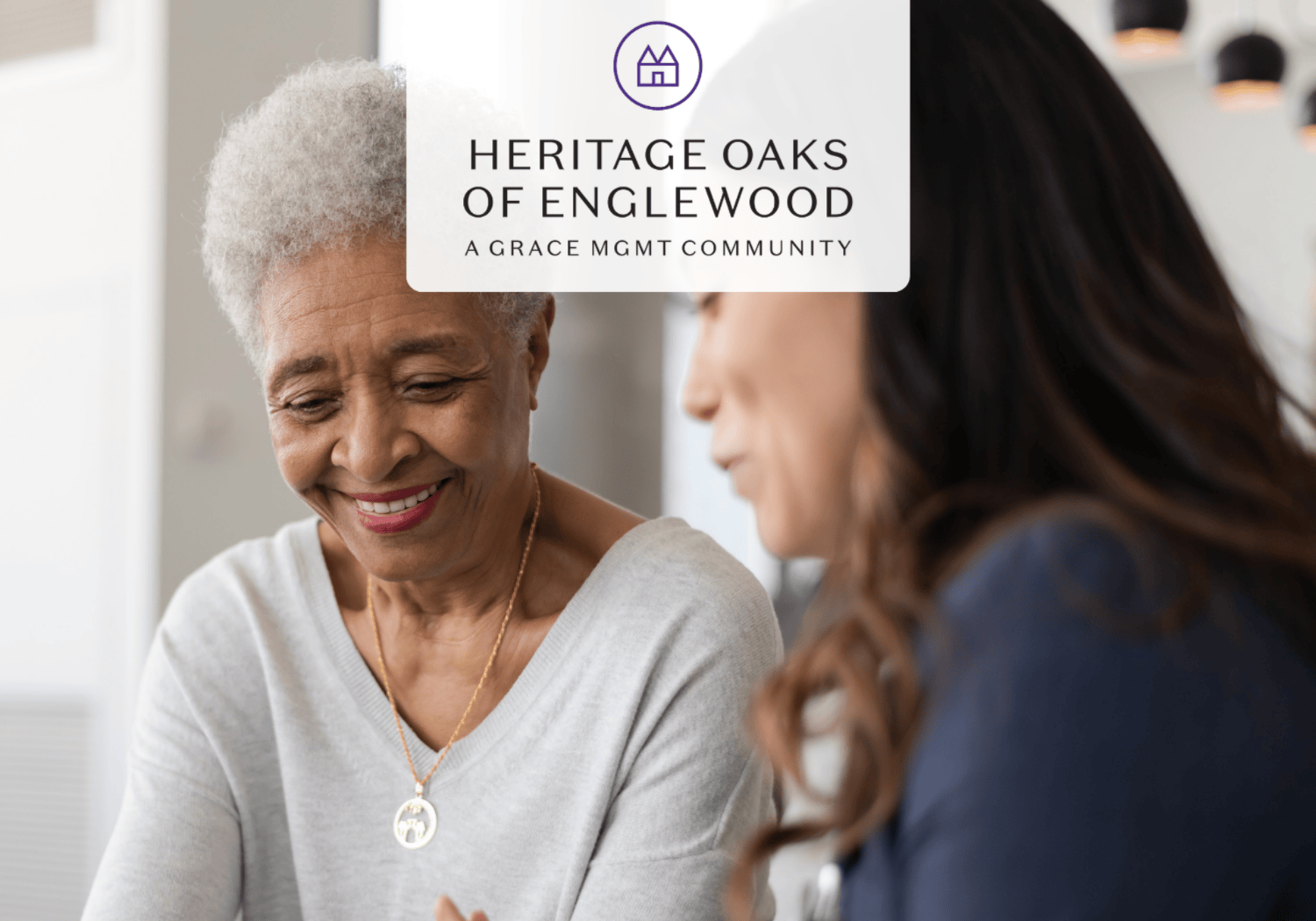 Older African American woman smiles while talking with a younger woman in a bright room; Heritage Oaks of Englewood logo overlays the scene.