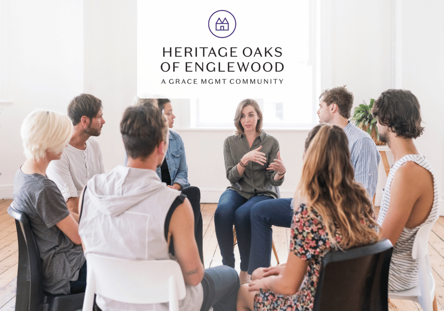 Group of adults seated in a circle in a bright room, a woman speaks in the center; the Heritage Oaks of Englewood logo is above the group.