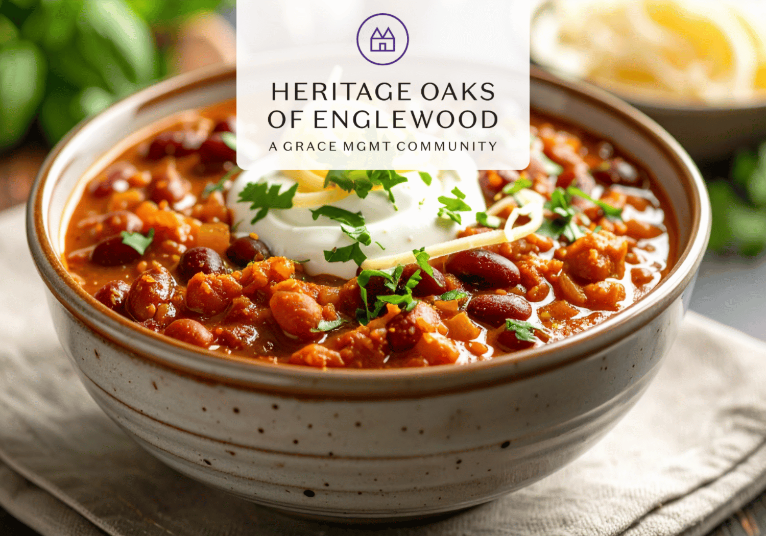 Creamy chili with beans in a ceramic bowl, topped with sour cream and parsley, with Heritage Oaks of Englewood logo overlay.
