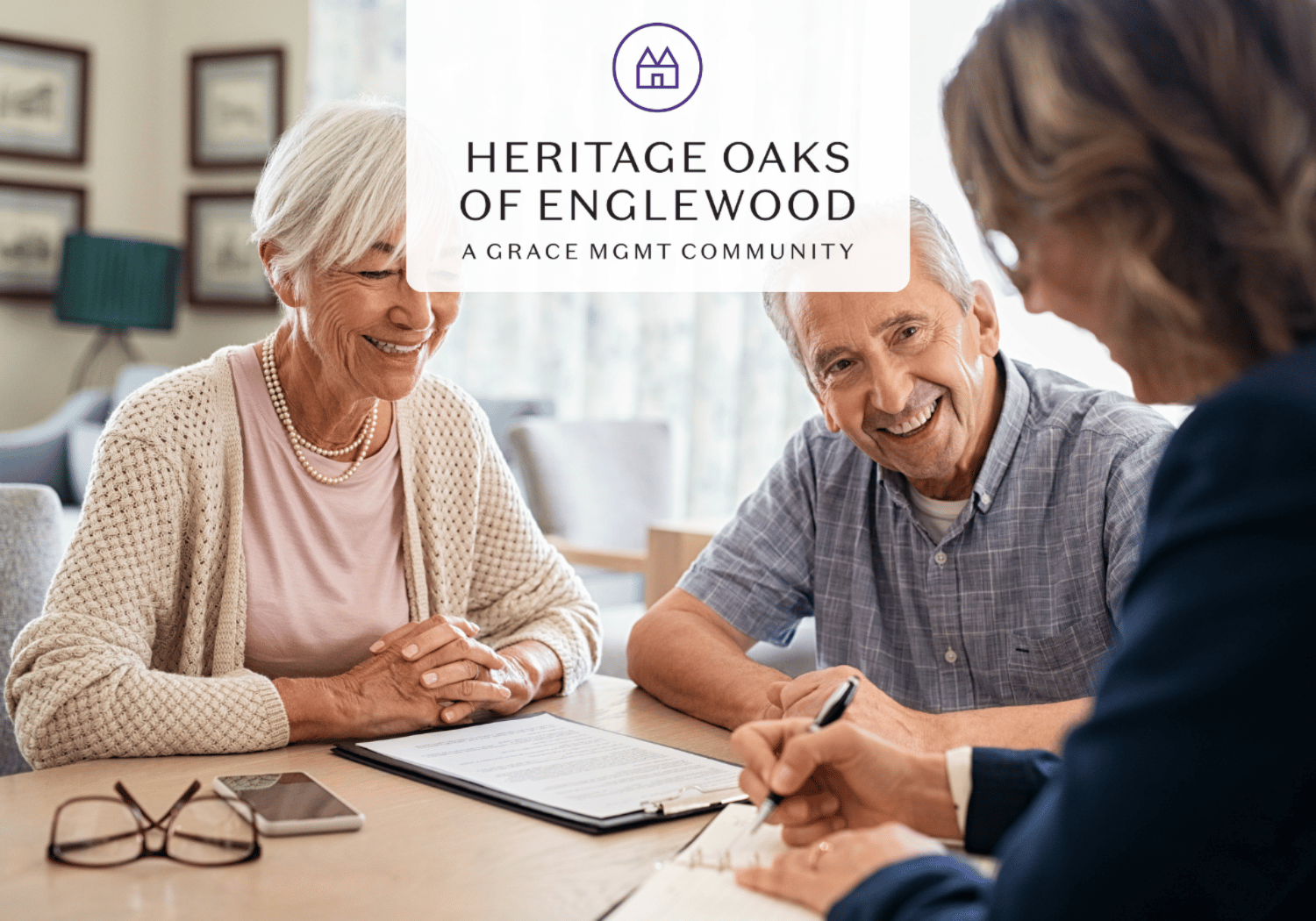 Elderly couple smiles at a staff member while signing paperwork, with Heritage Oaks of Englewood logo above them (a Grace MGMT Community).