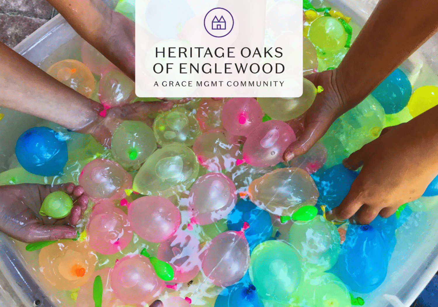 Children's hands reach for colorful water balloons in a tub, with a Heritage Oaks of Englewood logo overlay on top (community image).