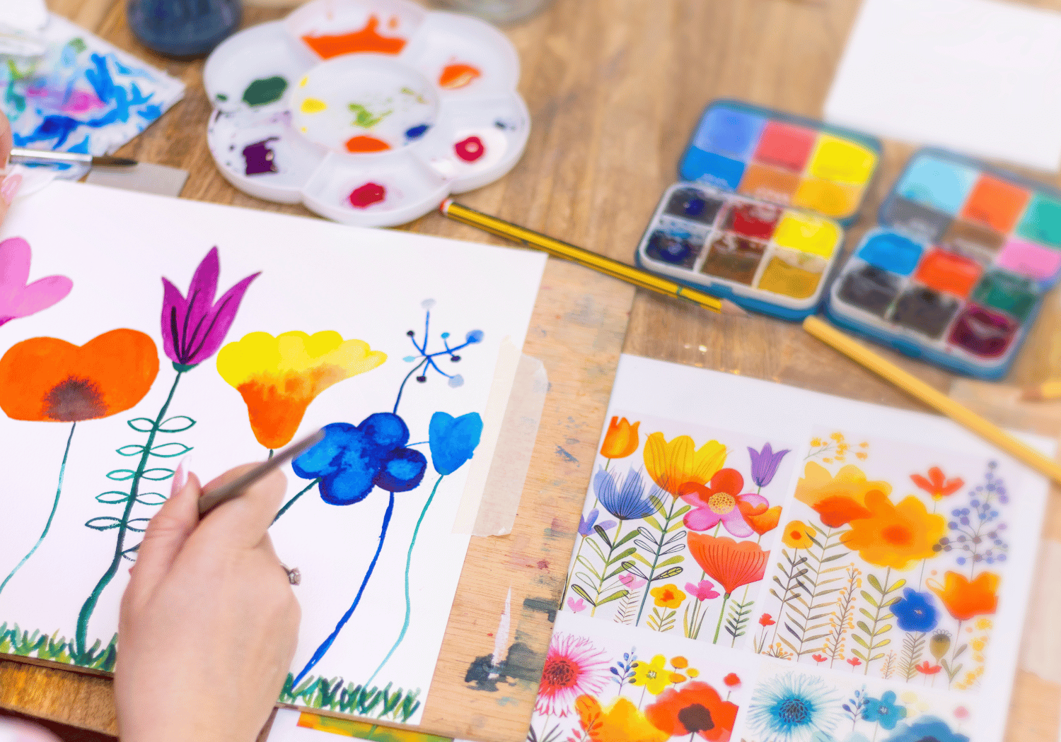 Hands painting bright watercolor flowers on white paper with a palette and brushes nearby on a wooden table.